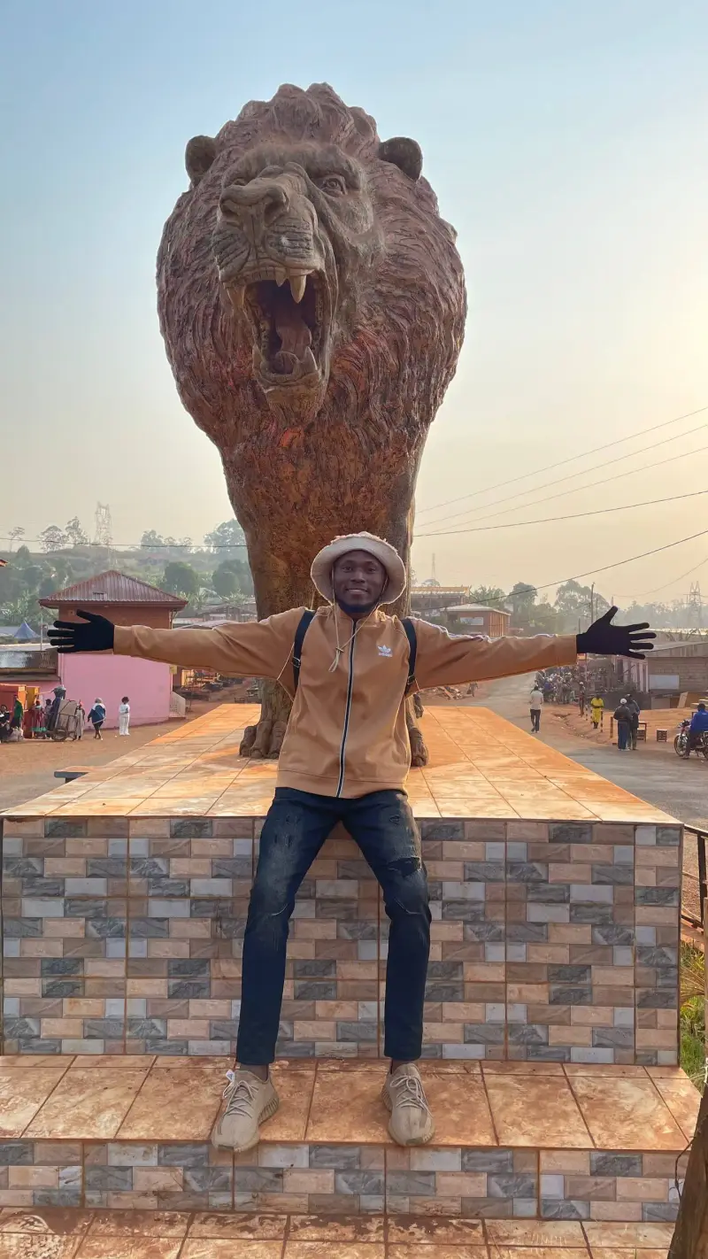 Njoh Simplice Junior standing arms outstretched in front of a monumental lion statue in Cameroon